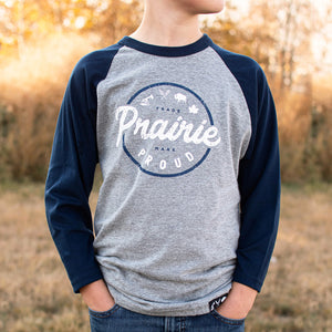 A child wearing a heather grey and navy raglan t-shirt with text 'Prairie Proud' printed on the front.