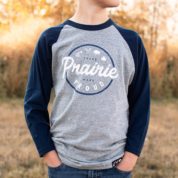 A child wearing a heather grey and navy raglan t-shirt with text 'Prairie Proud' printed on the front.
