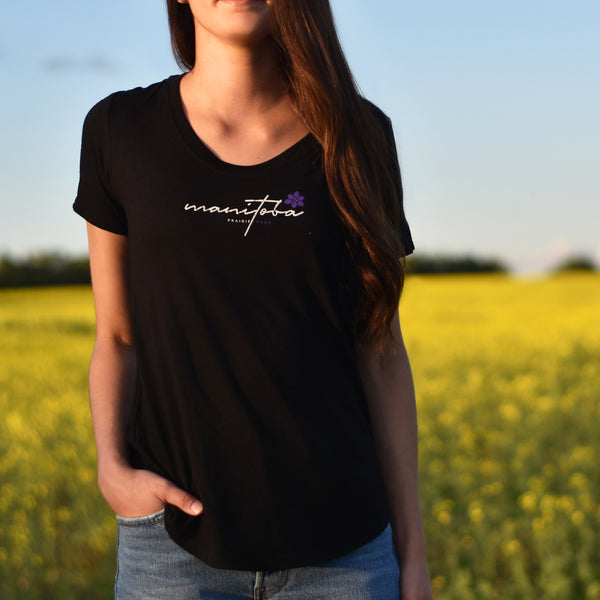A woman standing in a field wearing a black scoop neck t-shirt with the word 'Manitoba' written on it and a small purple flower.