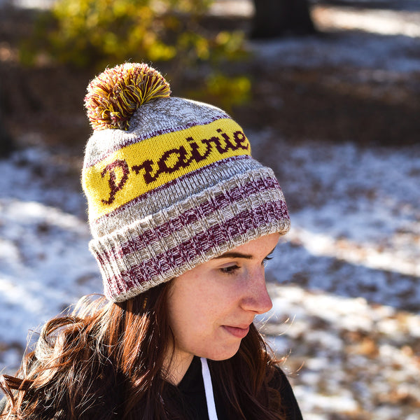 A person wearing a heather tan and gold knit hat with the text 'Prairie proud', accompanied by a pom-pom.