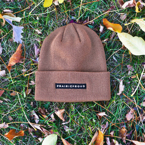 A caramel-colored beanie laid on a grassy surface with leaves around it, featuring a cuff with embroidery.