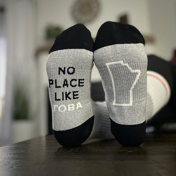 A pair of black and grey crew socks with text 'NO PLACE LIKE TOBA' on the right sock, and the province shape on the left.