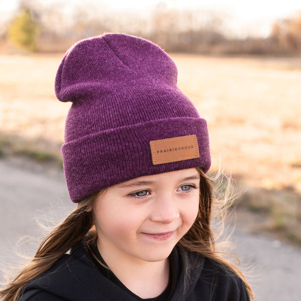 A young child wearing a heather blackberry coloured beanie with a leather patch on the cuff.