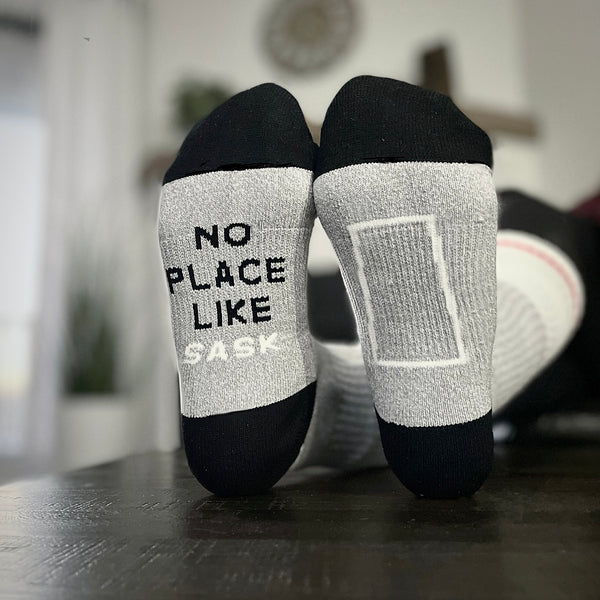 A pair of black and grey crew socks with text 'NO PLACE LIKE SASK' on the right sock, and the province shape on the left.
