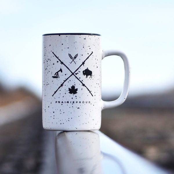 A white ceramic mug with black speckles and a design featuring crossed barb wire with various symbols, along with the text 'Prairie Proud'.
