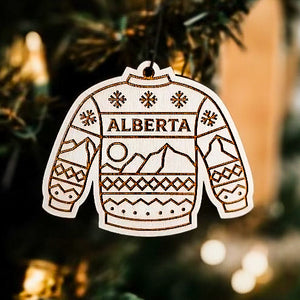 Christmas ornament in the shape of a sweater with the text Alberta and a pattern of mountains and snowflakes. 