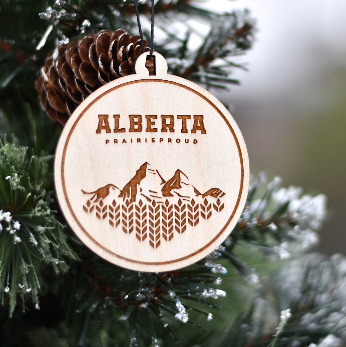 A circular wooden ornament with the engraved design of mountains and the text 'Alberta Prairie Proud' on a Christmas tree.