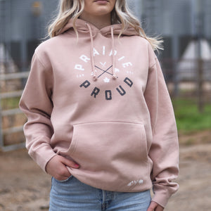 A woman wearing a dusty pink hoodie featuring a 'Prairie Proud' circular text logo in a farmyard setting