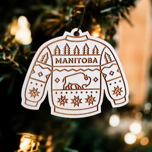 A wooden ornament in the shape of a sweater hanging on a Christmas tree, with an engraved design featuring a buffalo, trees, and the word 'MANITOBA'.