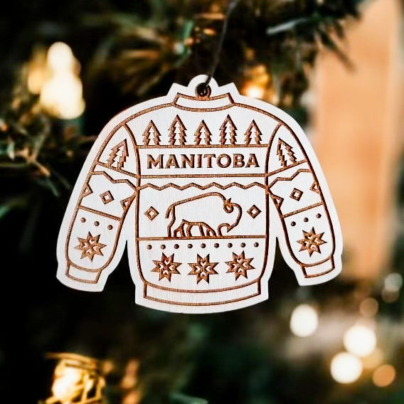 A wooden ornament in the shape of a sweater hanging on a Christmas tree, with an engraved design featuring a buffalo, trees, and the word 'MANITOBA'.