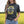 A woman wearing a dark charcoal gray graphic t-shirt with yellow and white canola floral designs and the text 'PRAIRIE PROUD'. She is wearing blue jeans and a black belt, standing in a field of yellow canola flowers under a bright sky.