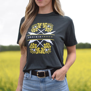 A woman wearing a dark charcoal gray graphic t-shirt with yellow and white canola floral designs and the text 'PRAIRIE PROUD'. She is wearing blue jeans and a black belt, standing in a field of yellow canola flowers under a bright sky.