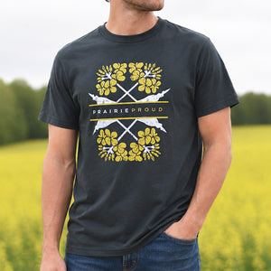 A man wearing a dark charcoal gray t-shirt with a floral yellow canola design and white 'Prairie Proud' graphic, standing in a blurred yellow canola field.
