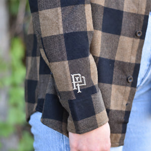 Close-up of a person wearing a brown and black buffalo plaid flannel shirt with a white embroidered 'PP' monogram logo on the cuff, paired with light blue jeans.