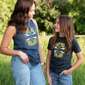 A woman and a young girl standing outdoors in a grassy field, both wearing matching navy blue graphic t-shirts with a yellow and white canola design and the text 'PRAIRIE PROUD".