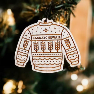 A wooden ornament in the shape of a sweater with "Saskatchewan' written on it hanging on a Christmas tree, with engraved design details.