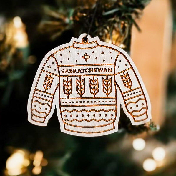 A wooden ornament in the shape of a sweater with "Saskatchewan' written on it hanging on a Christmas tree, with engraved design details.