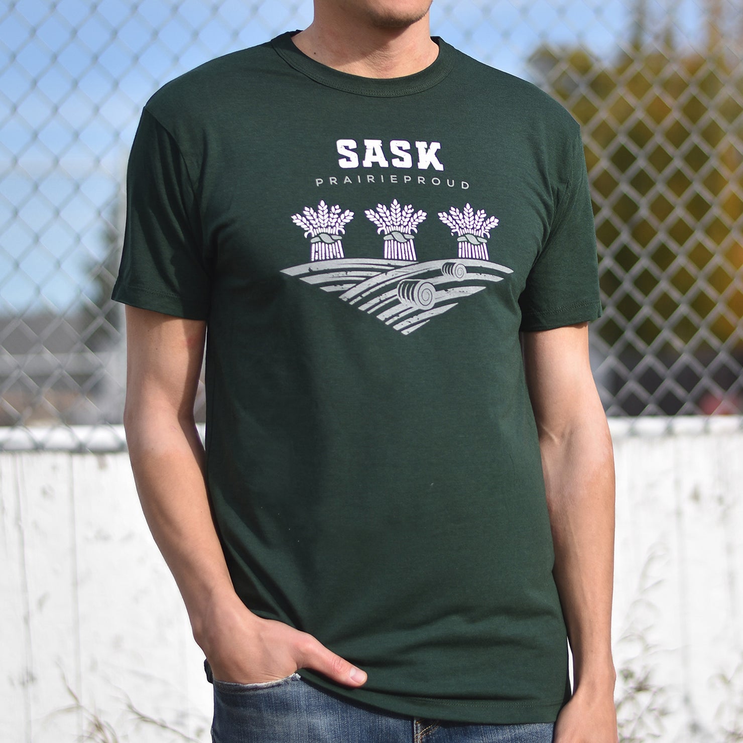 A person wearing a green crew t-shirt with 'SASK' text and a graphic on it standing in front of a chain-link fence.
